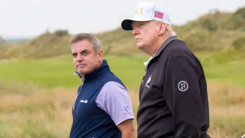 PA Media Dwóch mężczyzn idą po wodach pola golfowego. Prezydent Trump ma na sobie czarny wiatr z pół zip i białą czapkę baseballową z USA w złotych literach. Ma również Saltire z boku. Paul McGinley ma brązowe/ siwe włosy w bocznej szopie. Ma na sobie fioletową koszulę polo i bez rękawów niebieski zamek błyskawiczny. Obaj mężczyźni patrzą w lewo.