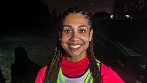 Bianca stands outdoors at night on a grassy area, wearing a bright pink long-sleeve top and a reflective safety vest with a glowing green light attached to the front. She has long braided hair and is posing with one hand on the hip.