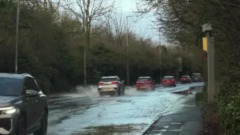 Cars drive along a busy road covered in what appears to be around two inches of water.