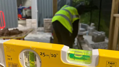 Megan Paterson A yellow spirit level is in the foreground of the picture, with the level bubble in yellow fluid to the right of the frame. In the background a teenage boy wearing a high-vis waistcoat is building a wall. The boy is out of focus with his back to the camera.