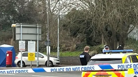 A close-up picture of a blue and white police cordon which says "police do not cross". There is also a police car and others in the scene and a blue police tent, postbox and car in the background. 