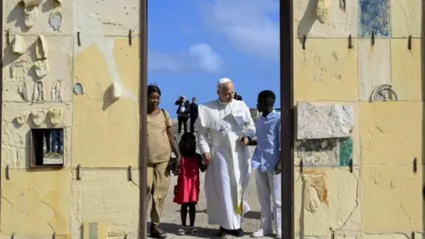 Vatican Media Pope Leo holds hands with a girl wearing a red dress and a man in blue shirt and white trousers. A women in a beige outfit stands alongside them at a doorway. Behind them are blue skies and men dressed in black