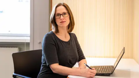 Royal College of Nursing A woman with a brown bob and glasses sits at a desk next to an open laptop. She is wearing a dark grey long sleeved top and looks directly into the camera. 