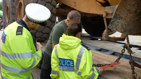 BBC/Gary Darfield A man and a woman in high vis yellow jackets with a blue police badge on the back stand with a man in a green tshirt and navy cargo trousers in front of trailer carrying a vehicle with large tyres. 