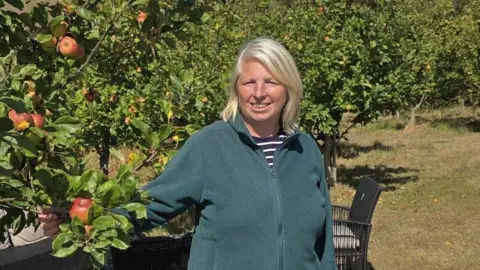 Cuckoo Valley Cider Lorraine O'Connor is standing in an orchard next to apple trees. She's smiling at the camera and is holding one of the branches which has a red apple on it. 
