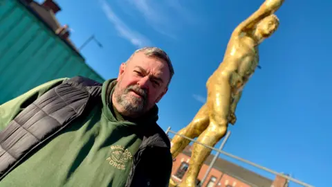 A man in a green Gerentaya hoodie has short hair on his head and the dust-effect is raised in the air. It was born with a male but it was removed to the background in this shot. The blue sky and rooftops can be seen behind the big statue. 