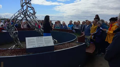 A choir singing standing outside. They are gathered around in a semi-circle.