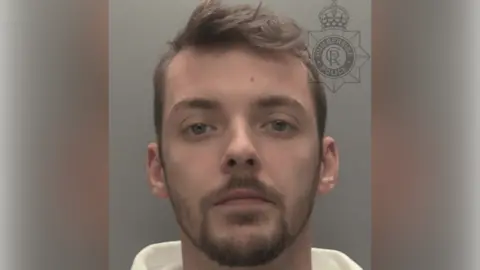 A police custody shot of Jacob Agnew. He is wearing a white hoodie and has short brown hair and short brown facial hair. He is looking at the camera. The wall behind him is grey.