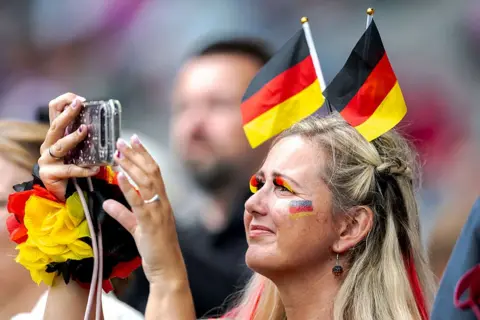 Getty Images A German fan takes a picture with her phone. She is wearing eyelashes in the colours of the German flag and two small flags tucked into her hair 