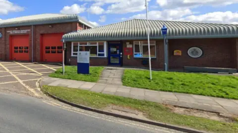 A fire station pictured from the road. It is made up of two single-storey buildings. The taller one, on the left, includes two traditional red garage style shutters behind which fire engines are parked. Each shutter has six rectangular windows in two rows of three. The shorter building, on the right, has a blue door with windows either side and a longer expanse of brick with an  old style police lamppost outside. Each building has a grey pyramid-shaped corrugated roof. 