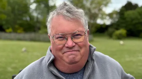 Jodie Halford/BBC Mark Dutton has grey hair and glasses with silver rims. He is wearing a grey jumper in front of a grassed area outdoors.
