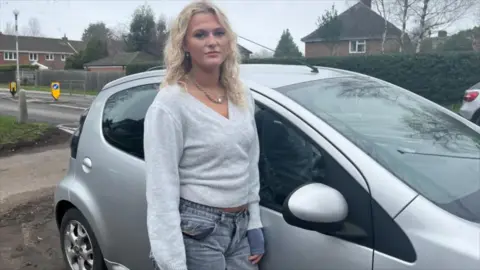 A woman wearing a grey cardigan and jeans looks at the camera whilst standing next to her silver car.