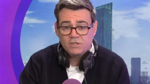 Andy Burnham on radio manchester with purple background. He has dark rimmed glasses a black jacket and white T-shirt