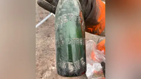 A green bottle is held up to the camera. It has the words High Street Crewe embossed on it