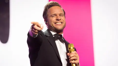 Olly Murs performs onstage - he is holding a grin microphone in one hand, grinning and pointing  with his other hand