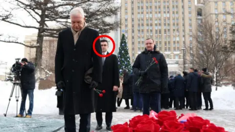 Telegram Russian Deputy Foreign Minister Alexander Grushko, a man with white hair, wearing a dark overcoat and a scarf, stands with his head bowed, in front of a display of red flowers seemingly laid as a memorial. Behind him stands Evgeny Lyukshin, a young man with dark hair and glasses, wearing a dark overcoat and tie, holding a couple of red flowers. There is a crowd of people further behind in what appears to be a snowy Moscow public square them and a man is filming the event with a camera on a tripod.