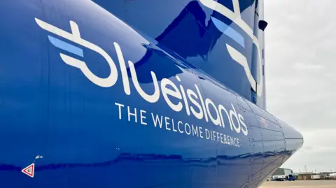The side of a Blue Islands aircraft parked  on the runway at Jersey Airport. It is  bright blue and has the company name in white on the side, with the words The Welcome Difference below. they are also in white. A bus is in the far distance.