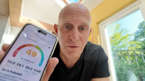 A man holds up a mobile phone showing declining air quality within his home as he cooks and cleans.