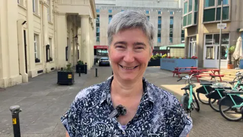 Abigail Dombey/BBC A woman with grey hair is wearing a blue shirt with black and white flower prints. She is standing on a street.
