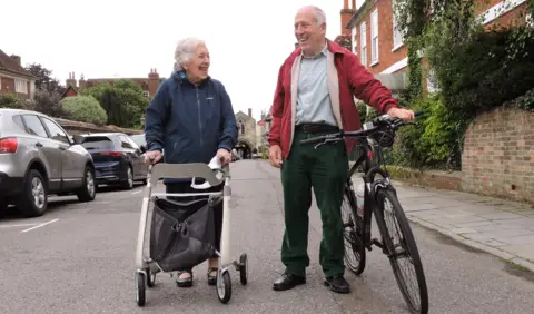 Phyll with a walker makes her way down a road, laughing with a man pushing his bike