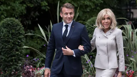 French President Getie Emmanuel Macron and First Lady Bridget Macron arrive on Downing Rate in July. The picture shows a couple who walked his hand in his hand. Mr. Macron is dressed in a dark -headed suit, a white shirt and a dark tie. Mrs. Macron dresses in a light-colored, double-breasted suit. They are surrounded by greens, including shrubs and plants with long leaves