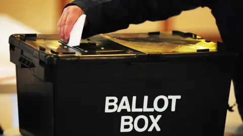 PA Media A black plastic box with the words "Ballot Box" printed on it in white. A person, who is wearing a black coat, is placing a folded piece of paper through the slit in the top.