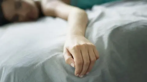 Getty Images A stock image showing a woman lying face-down on a bed with pale blue linen sheets. Her hand is limp in the foreground and she has her eyes closed. Most of her body is blurred or not visible.