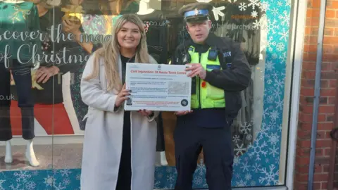 Huntingdonhire District Council Georgia Patterson, community safety partnership delivery officer and PCSO Daniel Grant, standing outside a shop front holding a sign. Georgia is to the left and wearing a light coat, and has long fair hair, PCSO Daniel Grant, is wearing full police uniform, with a high viz jacket. 