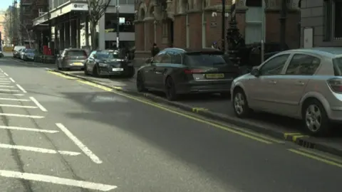 There are eight vehicles parked on the footpath next to a double yellow line. The cars are a mix of colours including silver, dark grey and black. There red brick and white buildings along the side of the street.