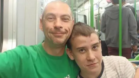 Colin and Rhys seated together on an underground train. Colin wears a bright green t-shirt bearing a Fox logo, and smiles at the camera. He has one arm around his son's shoulder and one arm on the carriage window sill. Rhys is wearing white and grey stripped t-shirt and has his head on his father's shoulder. 
