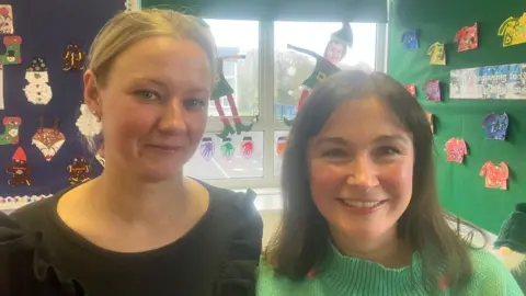 Gillian has a green jumper and brown hair and Gintare has a black top and blond hair. They are in a classroom. 