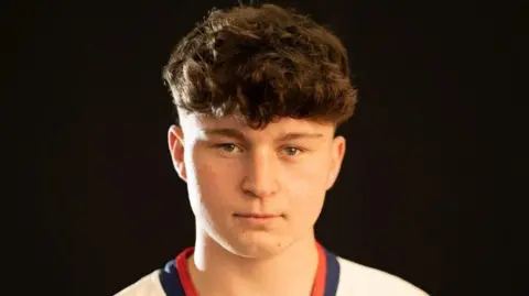 JACOB MANSELL A close-up of the face of young man with curly hair. He wears a white top with a blue and red collar.