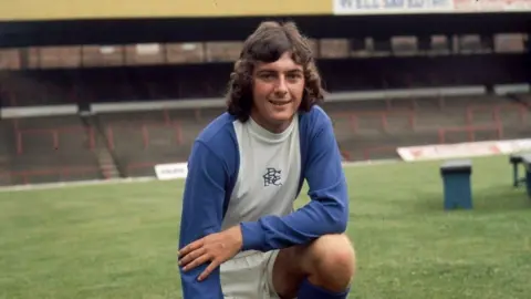 A man kneels on a football pitch. He has chin-length brown hair and he is wearing a blue and white sports kit. The football ground's seats can be seen in in the background.