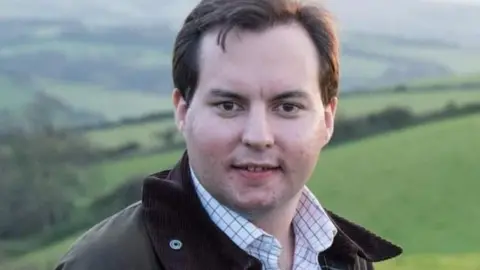 Somerset Council Theo Butt-Philip is seen from his shoulders up in front of a hilly background. He is wearing a leather jacket and a white shirt. He is smiling at the camera.