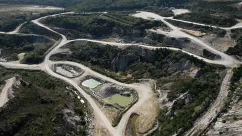 Cornish Lithium  Ariel view of Trelavour pit. There are green areas amongst grey stone paths leading to three pits. The pit on the left has blue coloured water inside, the one in the middle has blue water and the one on the right has green water. They are surrounded by cliff faces covered with trees.