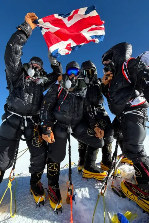 Sandro Gromen A group of four mountaineers stands on a snowy summit under a clear blue sky, wearing heavy cold-weather gear including thick black insulated suits, gloves, and crampons. Several have face masks and goggles covering much of their faces. One mountaineer holds a Union Jack flag above the group.