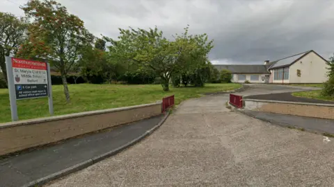 A Google Streetview screenshot of the former St Mary's Church of England Middle School in Belford, Northumberland. A concrete driveway leads to an L-shaped cream building with large windows. There is a sign with the name of the school at the side of the road.