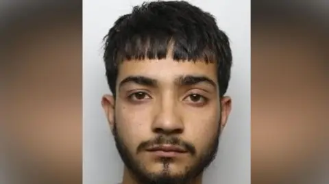 A man with a short black beard and goatee. He has short straight black hair cut straight across is forehead. He has an unhappy expression.