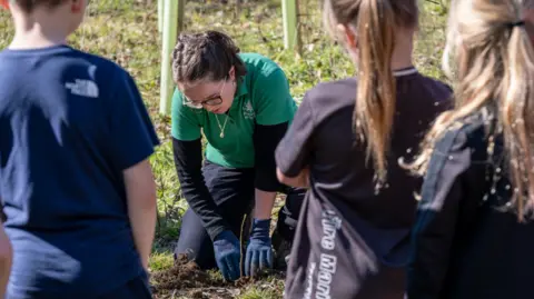 Alison Whaley A young woman with tied back brown hair and glasses, who is wearing a green Forestry England polo shirt, plants a tree in the ground as primary school aged children watch on in the sunshine.