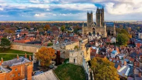 An aerial shot of part of Lincoln. The cathedral is on the far right and there are sand-coloured walls in the foreground both surrounded by redbrick buildings. Countryside and the Lincolnshire coast can be seen in the distance.