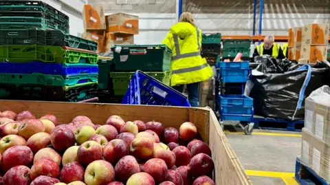 A box full of red apples is in the foreground, while volunteers in his vis jackets pack crates of food in the background.
