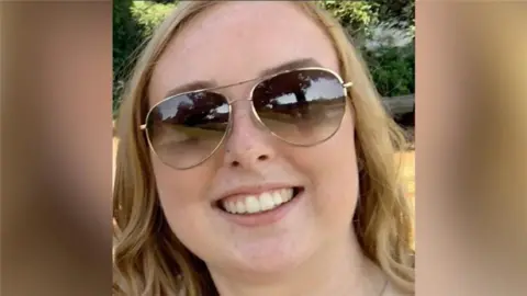 A photograph of Stephanie Irons, a 23-year-old white woman. Se is smiling, wearing aviator style sunglasses and has wavy hair