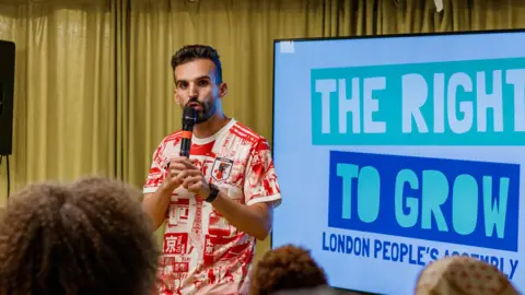 Tanya Vora Mete Coban speaking into a microphone next to a large blue and white presentation screen that reads "The Right to Grow London People's Assembly".