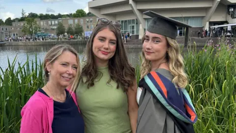 Hefina Everard Helen wearing a navy dress and pink cardigan, has blonde hair pinned up. She is standing with her daughters Hefina and Manon. Hefina is wearing a green dress and has dark hair, while Manon is wearing a graduation cap and gown. They are standing in front of a river, with reeds just behind them, and a concrete building with big windows and people in the background.