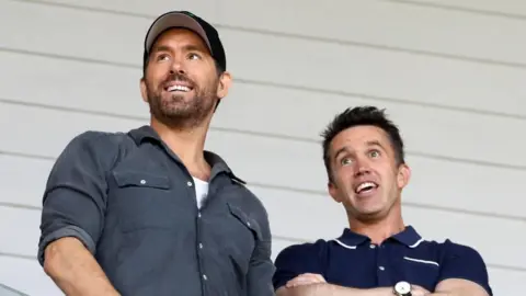 PA Media Ryan Reynolds in a cap and gray shirt stood next to Rob McElhenney in a blue polo shirt. They are both smiling and seem to be watching a soccer game.