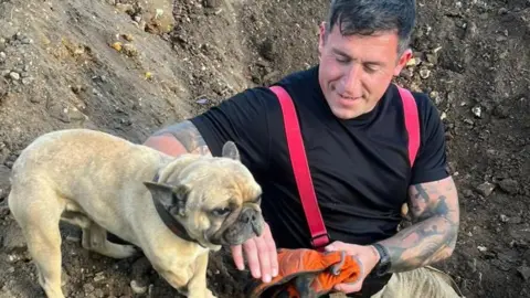 The French bulldog rests at the edge of the hole that was dug to rescue it. A firefighters sits slightly in the hole next to the dog and strokes it. 