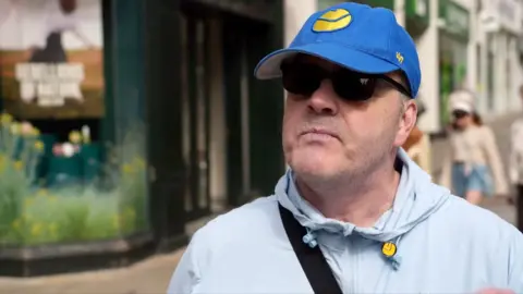 A man wearing a grey sports top with black glasses and a blue baseball cap with a yellow circular logo on the front