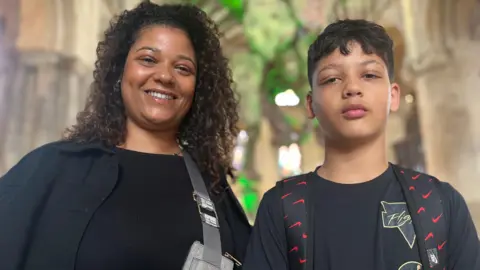 Malika, a woman in a black top, with curly brown hair, with a Eli, a boy in a black shirt. They are standing in front of the Titanosaur structure.