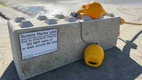 A white Government of Jersey warning signed screwed into a giant piece of stone. The signs advises people not to swim in or walk around the lake. They are two yellow buoys on top of the stone brick. 