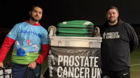 Tom Mansell Tom and Kyran are stood either side of the metal container the kit was carried in. It has a Prostate Cancer emblem on. Kyran is wearing an Alder Hey multi-coloured top and tracksuit bottoms. He has a beard and dark hair. Tom is wearing a black tracksuit with a Prostate Cancer logo on.